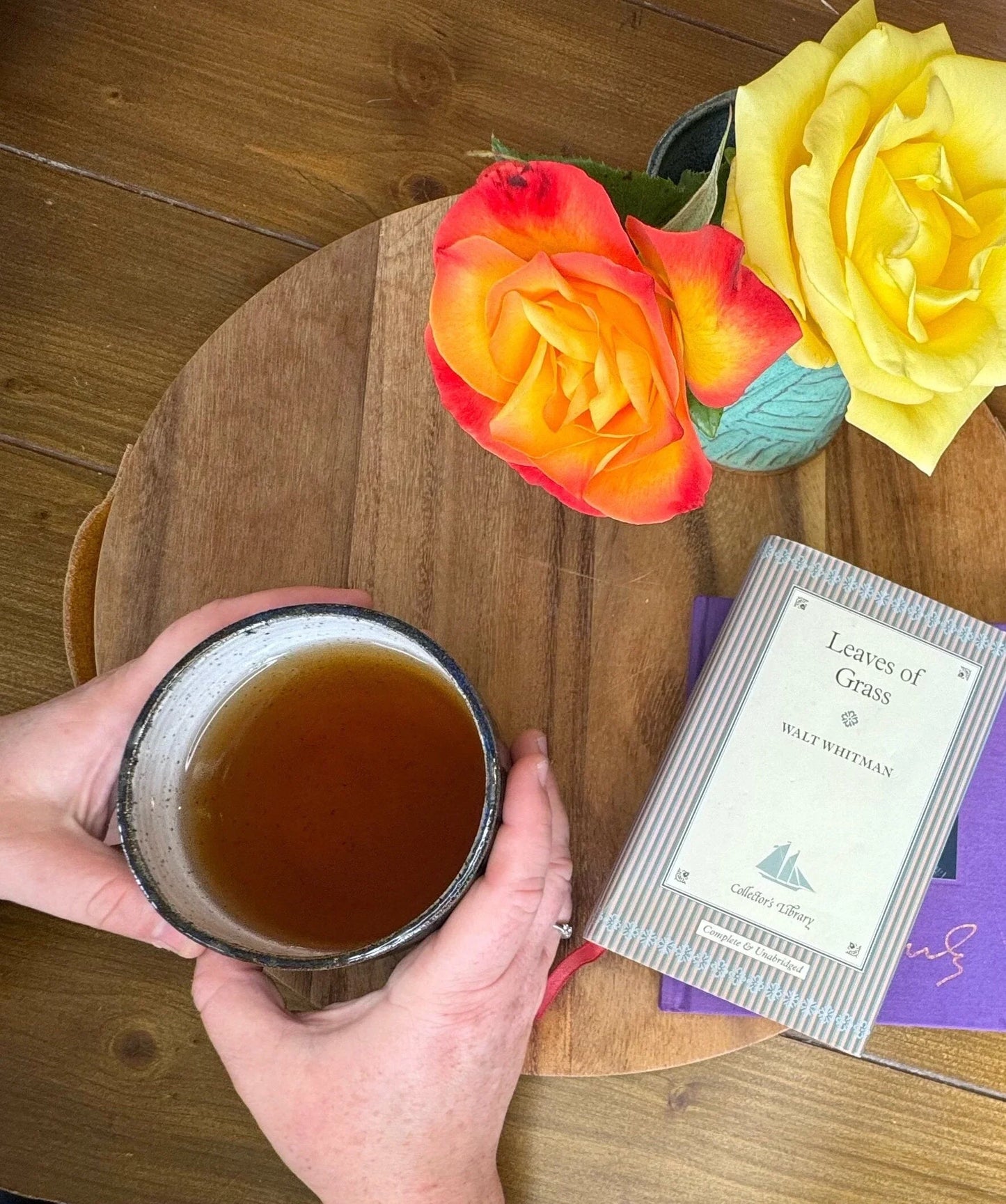 Hand holding a stoneware ceramic mug of tea beside roses in a vase and the book Leaves of Grass