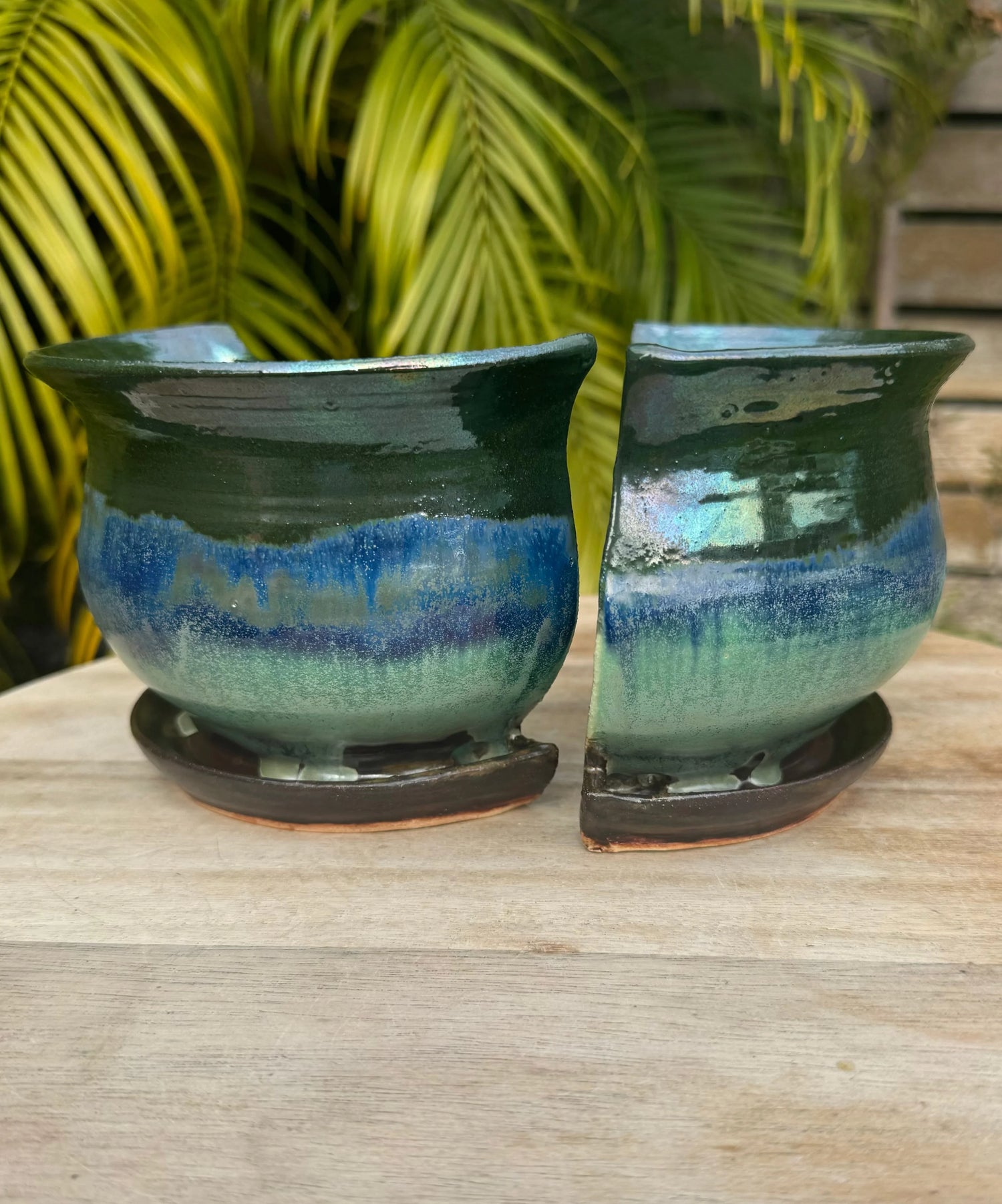 Handmade green and blue ceramic planter pots with drainage trays on a wooden surface, outdoor setting.