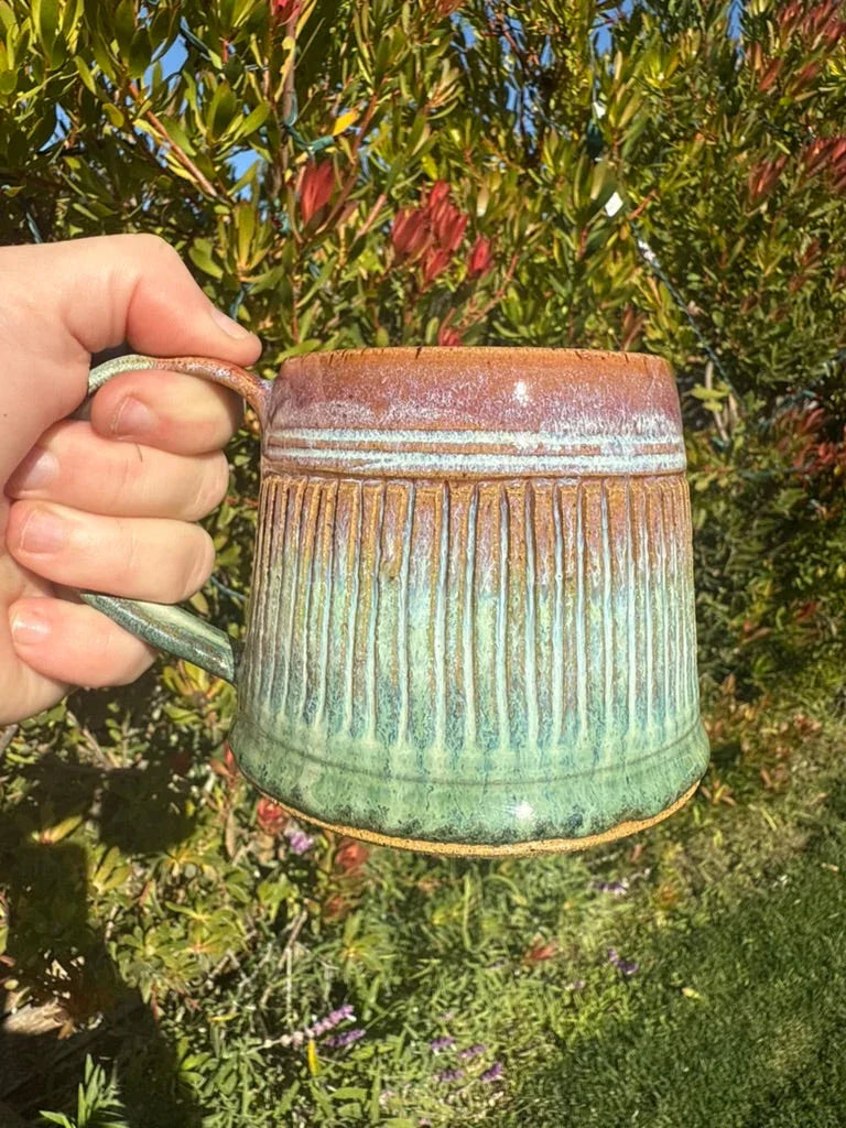Hand holding textured ceramic mug with green and brown glaze, outdoors in sunlight