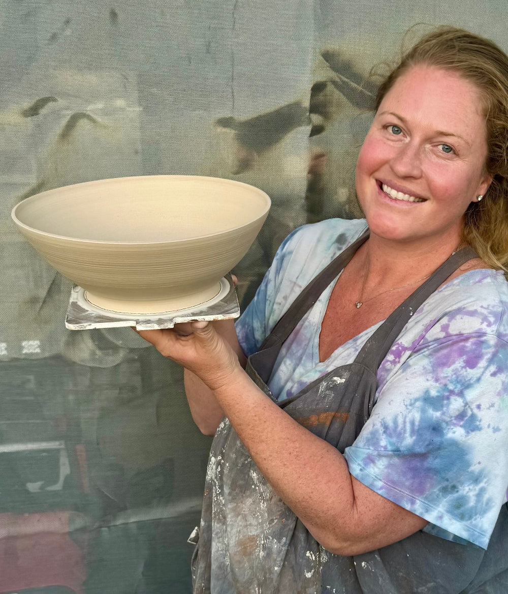 A smiling woman in a blue and purple tie-dye shirt and a stained apron holds a pale beige ceramic bowl on a small stand in front of a workshop backdrop.