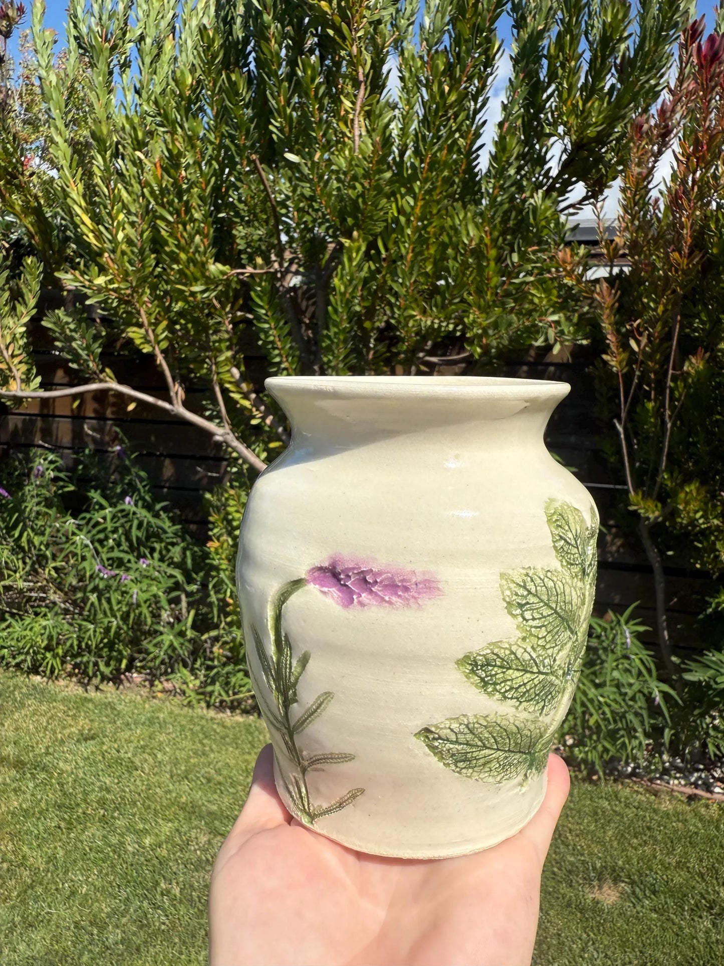Handmade stoneware vase with purple flower and green leaf design held outdoors